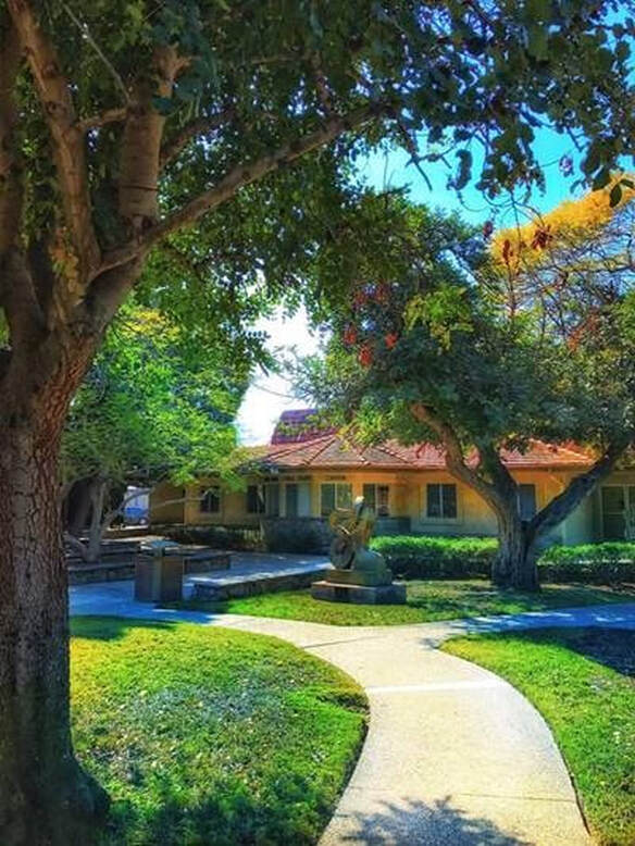 A cement path leads through landscaped grass and trees with a yellow building with a terracotta tile roof in the distance. An abstract bronze sculpture sits on the grass in front of the building.